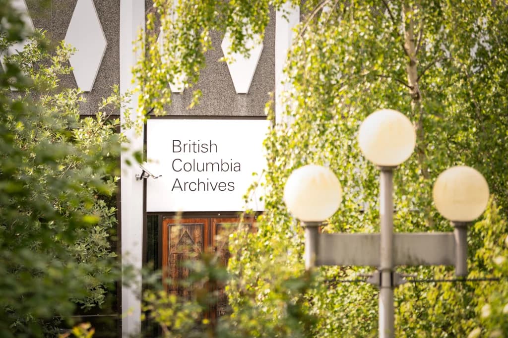 A photograph of the British Columbia Archives building exterior. It features a large sign with "British Columbia Archives" written in large black font on a white background, framed by a lot of greenery.