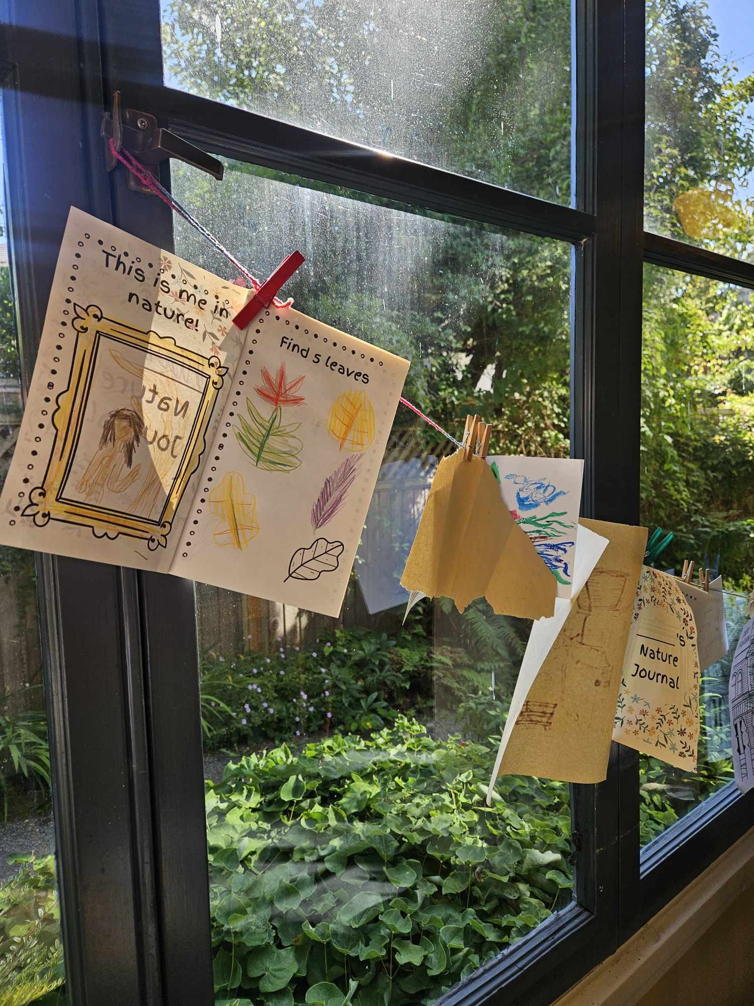 Hand-made children's nature journals pegged to a line across a window, with a green garden outside.